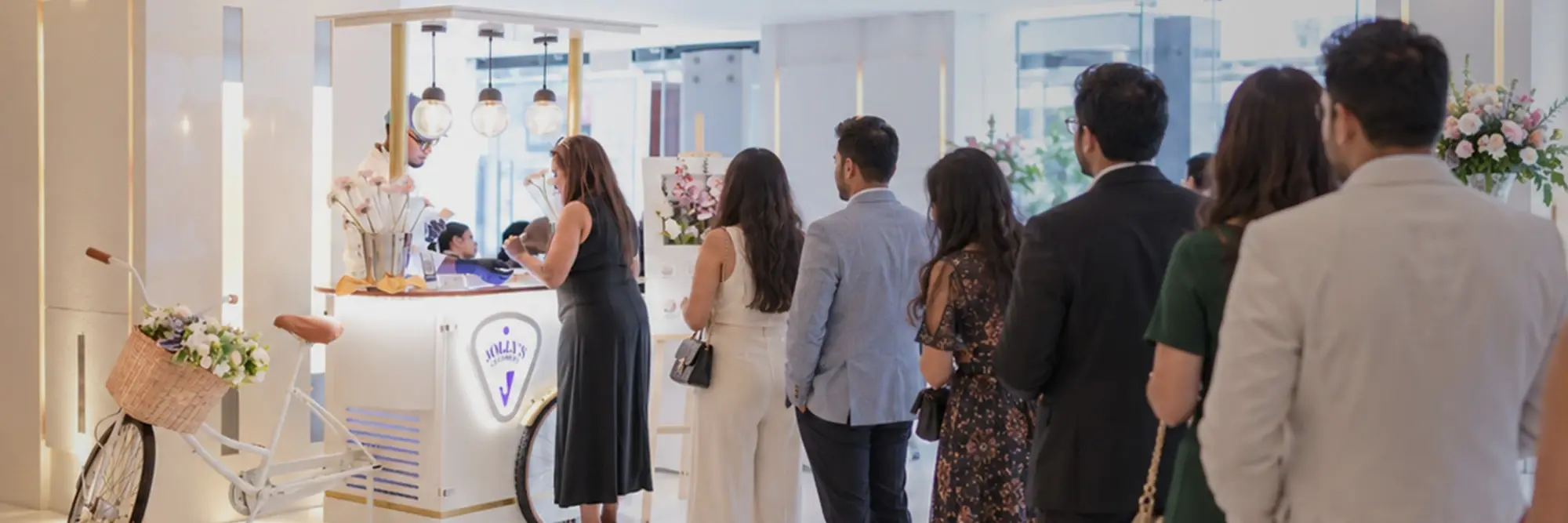 Guests in evening wear queueing at a Jolly's cart in a bright hotel lobby